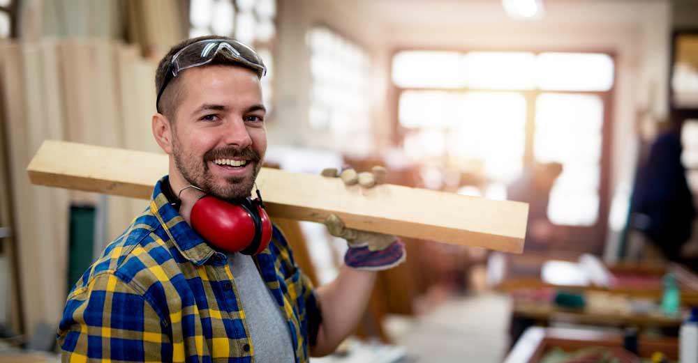 Smiling carpenter in a workshop holding a wooden plank on his shoulder, wearing safety goggles and ear protection, showcasing craftsmanship and DIY skills. Ideal for articles on woodworking techniques and safety measures in home improvement projects.
